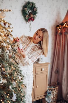A beautiful stylish woman in a white dress near the Christmas tree is smiling joyfully and wishes happiness.