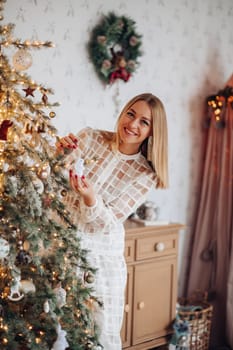 A beautiful stylish woman in a white dress near the Christmas tree is smiling joyfully and wishes happiness.