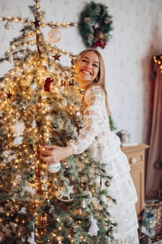A beautiful stylish woman in a white dress near the Christmas tree is smiling joyfully and wishes happiness.