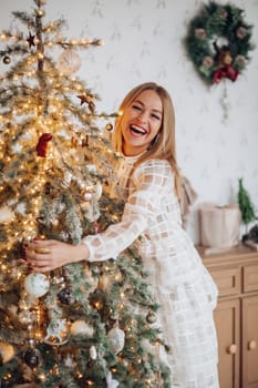 A beautiful stylish woman in a white dress near the Christmas tree is smiling joyfully and wishes happiness.