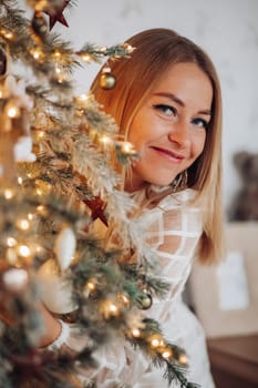 A beautiful stylish woman in a white dress near the Christmas tree is smiling joyfully and wishes happiness.