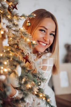 A beautiful stylish woman in a white dress near the Christmas tree is smiling joyfully and wishes happiness.