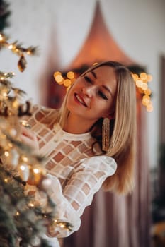 A beautiful stylish woman in a white dress near the Christmas tree is smiling joyfully and wishes happiness.