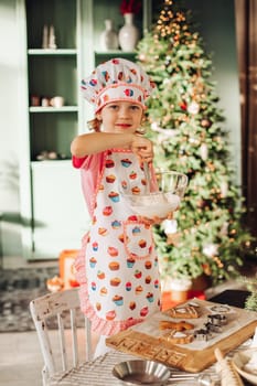 .a girl child in the kitchen with a Christmas tree is preparing or cooking ginger cookies for Christmas or New year