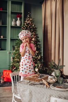 .a girl child in the kitchen with a Christmas tree is preparing or cooking ginger cookies for Christmas or New year