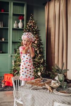 .a girl child in the kitchen with a Christmas tree is preparing or cooking ginger cookies for Christmas or New year