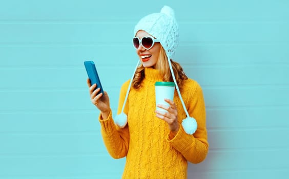 Winter portrait smiling young woman with phone and coffee cup wearing yellow knitted sweater, white hat, heart shaped sunglasses on blue background