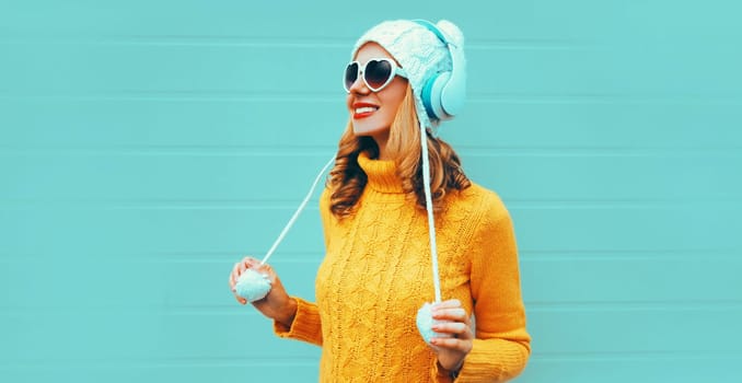 Winter portrait smiling young woman in headphones listening to music in yellow knitted sweater and white hat , heart shaped sunglasses on blue wall background