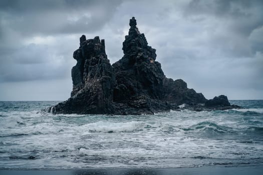 Dramatic black volcanic basalt formations and rocks amidst stormy ocean in cloudy weather.