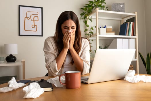 Sick woman working from home office. Caucasian female blowing nose with tissue while working with laptop. Wellness concept.