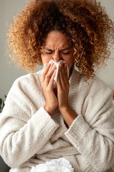 Vertical portrait of sick woman with the flu blowing nose with tissue paper at home. Multiracial hispanic female with a cold. Sickness or allergy concept.