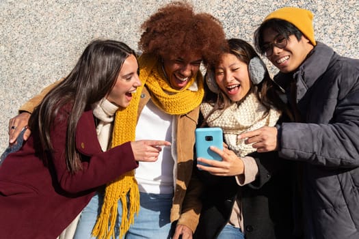 Happy multiracial college students friends looking at mobile phone together during sunny winter day. Young woman pointing at device screen. Technology and friendship concept.