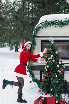 Young woman in santa costume decorates the Christmas tree at winter campsite getting ready for the new year. New year celebration concept