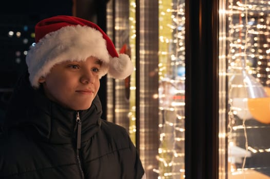 Boy in Santas hat looking and dreaming in illuminated shop window. Xmas presents holidays, or shopping on New Year or Christmas concept