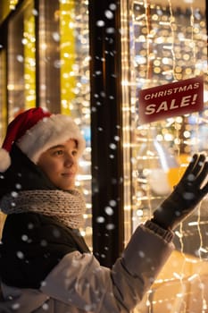 Boy in Santas hat looking and dreaming in illuminated shop window. Christmas holidays sales concept