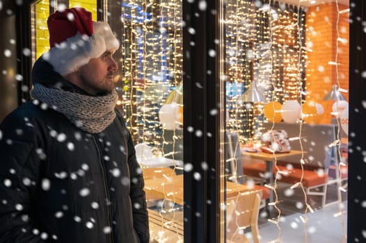 Man in Santas hat looking and dreaming in illuminated shop window. Christmas holidays sales concept