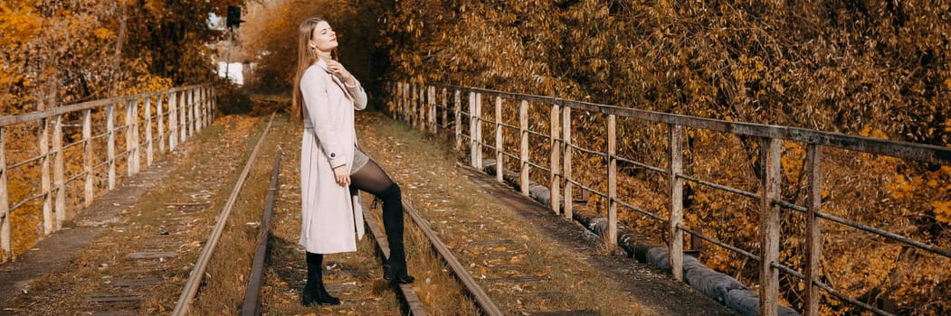 A beautiful long-haired woman walks through the autumn streets. Railway, autumn leaves, a woman in a light coat.