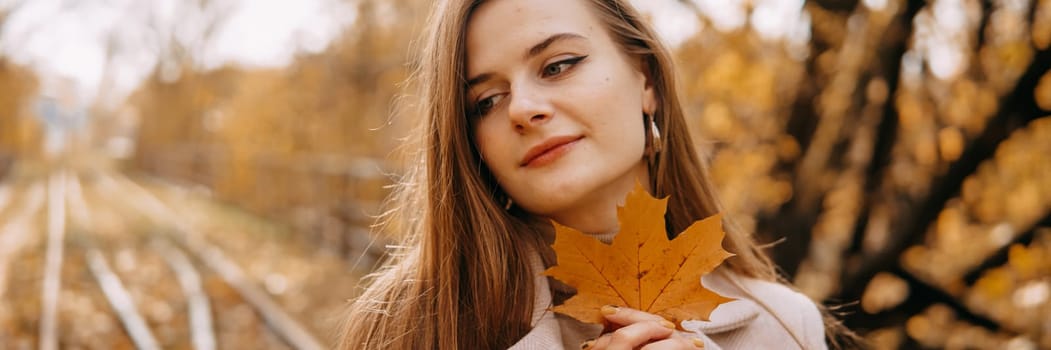 Portrait of a woman with an autumn maple leaf. Railway, autumn leaves, a young long-haired woman in a light coat coat, close-up.