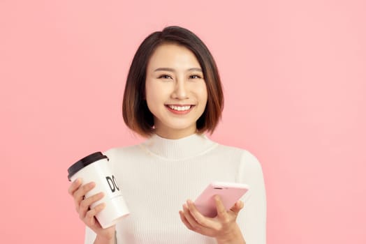 Close up portrait of young Asian woman holding coffee cup and smartphone over pink background.