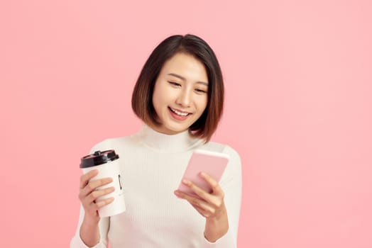 Attractive young Asian woman using mobile phone while holding coffee cup over pink background.