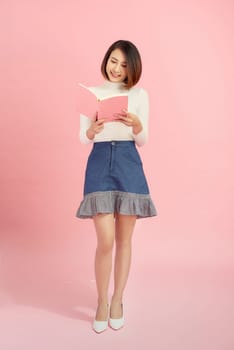 Young asian girl standing, reading interdting book.Isolated on pink background