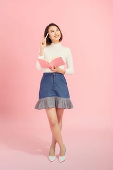 Full length portrait of a smiling beautiful Asian woman student holding book isolated on a pink background