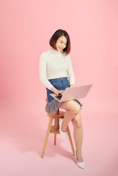 Young beautiful Asian businesswoman using laptop while sitting on chair. Isolated on pink background.