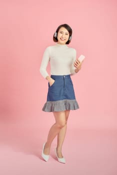 Full length portrait of a happy asian businesswoman listening to music with headphones while dancing isolated over pink background