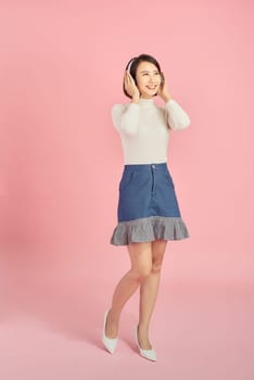 Full length portrait of a happy asian woman with headphones standing and holding mobile phone over pink background
