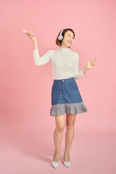 Full length portrait of a happy asian woman with headphones standing and holding mobile phone over pink background