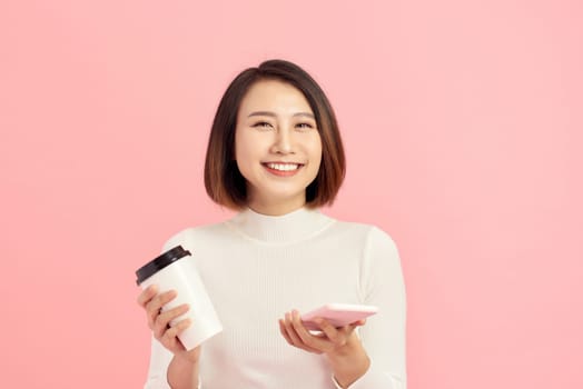 Close up portrait of young Asian woman holding coffee cup and smartphone over pink background.