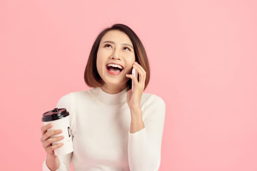 Portrait of cheerful young beautiful Asian woman listening smartphone while holding coffee cup over pink background.