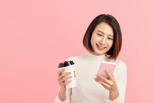 Attractive young Asian woman using mobile phone while holding coffee cup over pink background.
