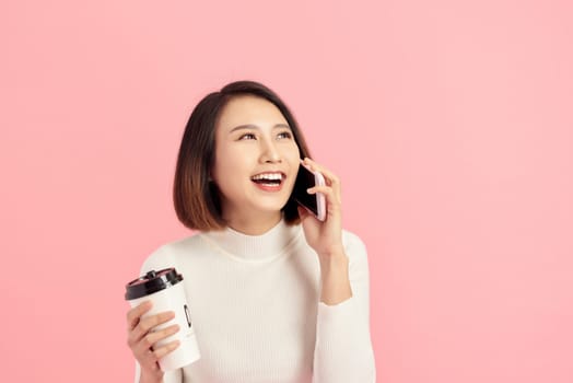 Portrait of cheerful young beautiful Asian woman listening smartphone while holding coffee cup over pink background.