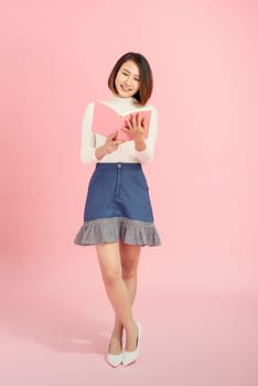 Full length portrait of a smiling beautiful Asian woman student holding book isolated on a pink background