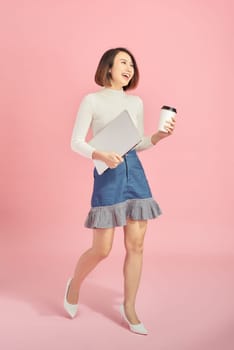 Young Asian woman holding laptop and coffee while walking over pink background.