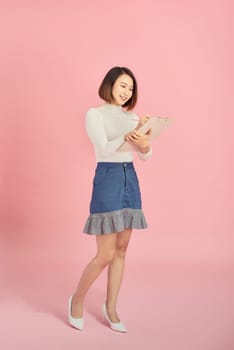 Full length of a smiling Asian woman standing isolated over pink background, holding notepad and pencil