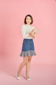 Full length of a smiling Asian woman standing isolated over pink background, holding notepad and pencil