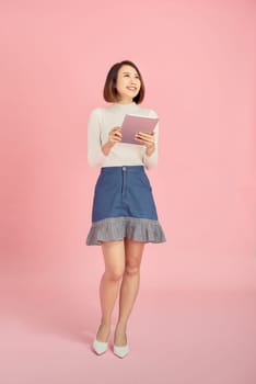 Young Asian woman reading book while standing over pink background.