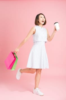 Smiling asian woman holding colorful shopping bags and coffee on raised hands isolated on pink
