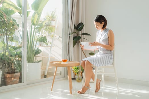 Woman relaxing and reading book near window in living room 