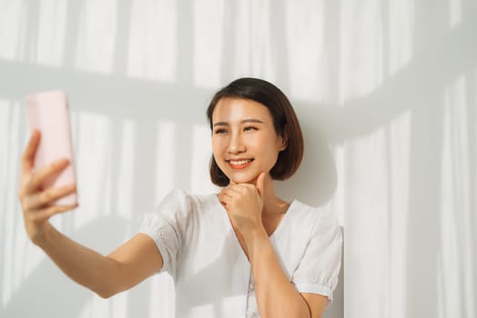 Friendly, tender young asian woman in casual outfit talking via internet video-call, holding smartphone taking selfie. 