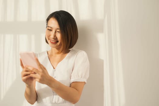 Beautiful young asian woman standing on white wall relax texting message on cellphone.