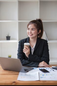 Photo of positive Asian businesswoman holds a cup of coffee imagines plan holiday in office..