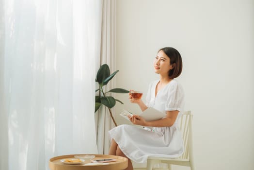 Young pretty woman sitting near window drinking tea and reading a book enjoys of rest