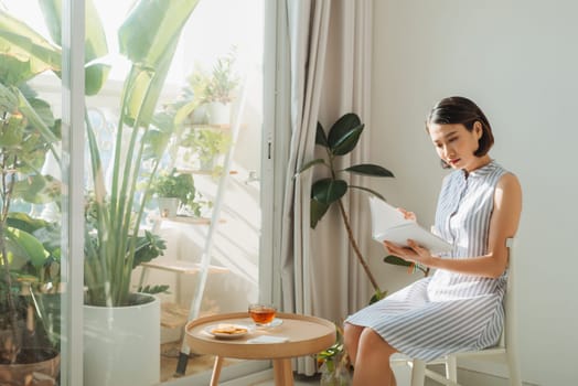 Beautiful young woman sitting by the window reading a book