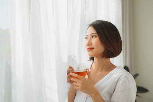 Portrait of a satisfied woman thinking looking at side holding a tea mug beside a window at home