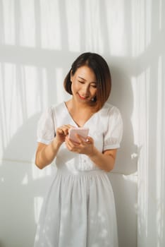 Content woman chatting on mobile phone while standing against white wall in sunset light