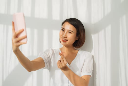 Sociable beautiful woman with asian appearance taking selfie or speaking on video call using cell phone isolated over white background