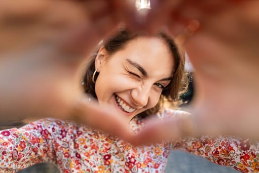 I love you. Portrait of Caucasian woman makes symbol of love, showing heart sign to camera, express romantic feelings, express sincere positive feelings. Charity, donation. Outdoors in city street